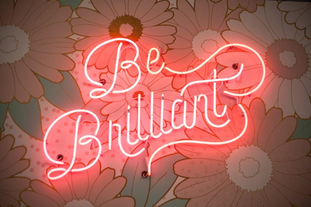 A glowing neon sign reading Be Brilliant against a floral backdrop creates a vibrant and motivational atmosphere.